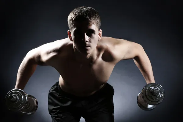 Handsome young muscular sportsman execute exercise with dumbbells on dark background — Stock Photo, Image