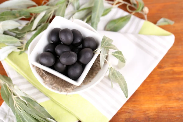 Оливки в миске с веткой на салфетке на деревянной доске на столе — стоковое фото