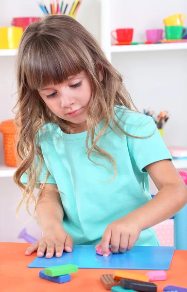 Pequena menina moldes de plasticina sentado à mesa no quarto em prateleiras fundo — Fotografia de Stock