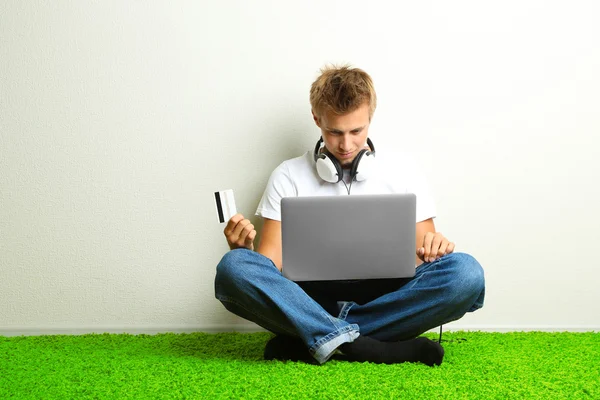 Jovem sentado no tapete com laptop e cartão de crédito, no fundo da parede cinza — Fotografia de Stock