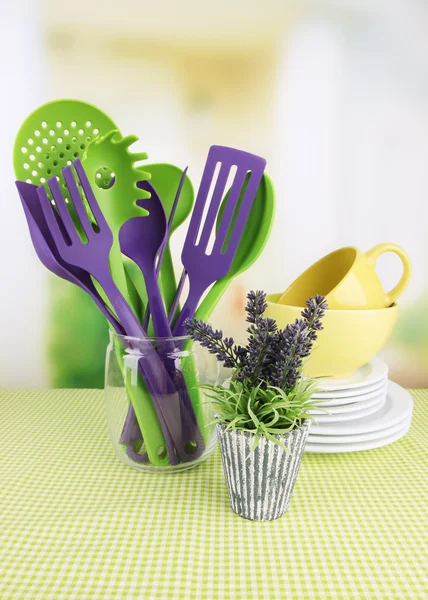 Plastic kitchen utensils in stand with clean dishes on tablecloth on bright background — Stock Photo, Image