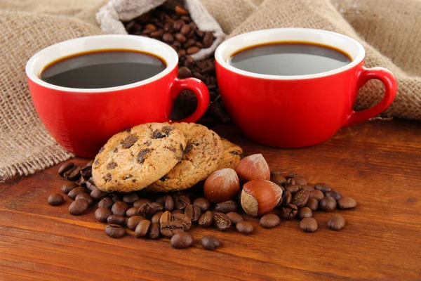 Tazas rojas de café fuerte con granos de café y galletas en la mesa sobre fondo de saco —  Fotos de Stock