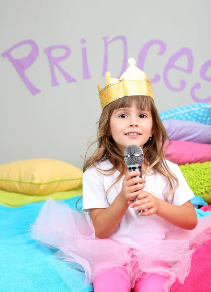 Μικρό κορίτσι που κάθεται στο κρεβάτι με το μικρόφωνο στο δωμάτιο σε φόντο γκρι τοίχων — Φωτογραφία Αρχείου