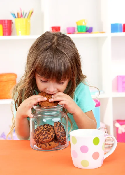 Little girl eating cookies sitting at table in room on shelves background — Stock Photo, Image