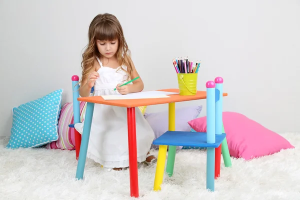 Niña dibuja sentado en la mesa en la habitación sobre fondo gris de la pared — Foto de Stock