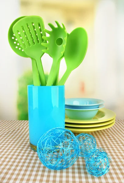 Plastic kitchen utensils in stand with clean dishes on tablecloth on bright background — Stock Photo, Image