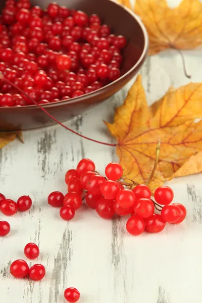 Красные ягоды вибурнума в миске и желтые листья на деревянном фоне — стоковое фото
