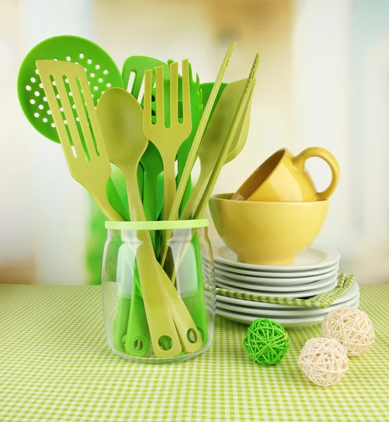 Utensílios de cozinha de plástico em stand com pratos limpos na toalha de mesa em fundo brilhante — Fotografia de Stock