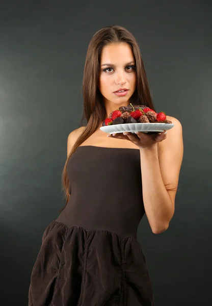 Portrait of beautiful young girl with chocolate sweets on plate on grey background — Stock Photo, Image