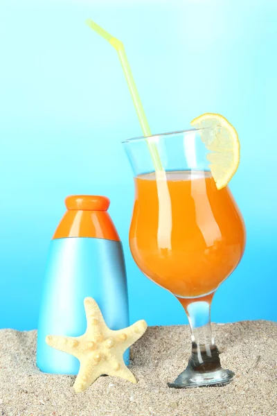 Beach cocktail and sunscreen in sand on blue background — Stock Photo, Image