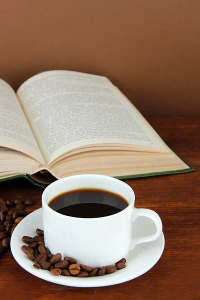 Kopp kaffe med kaffebönor och bok om träbord på brun bakgrund — Stockfoto