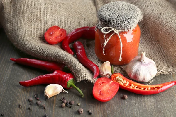 Composition avec sauce salsa dans un bocal en verre, piments rouges et ail, sur un sac, sur un fond en bois — Photo