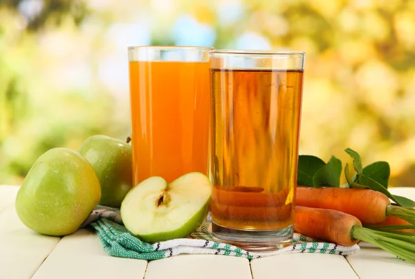 Glazen van SAP, appelen en wortelen op witte houten tafel, op groene achtergrond — Stockfoto