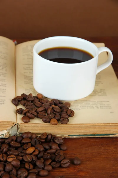 Cup of coffee with coffee beans and book on wooden table on brown background — Stock Photo, Image