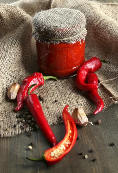 Composición con salsa en frasco de vidrio, pimientos rojos picantes y ajo, sobre tela de saco, sobre fondo de madera —  Fotos de Stock