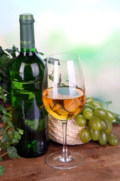 Ripe green and purple grapes in basket with wine on wooden table on bright background — Stock Photo, Image