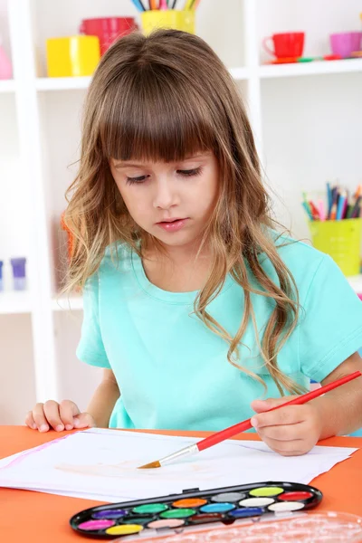 Маленькая девочка рисует сидя за столом в комнате на фоне полок — стоковое фото