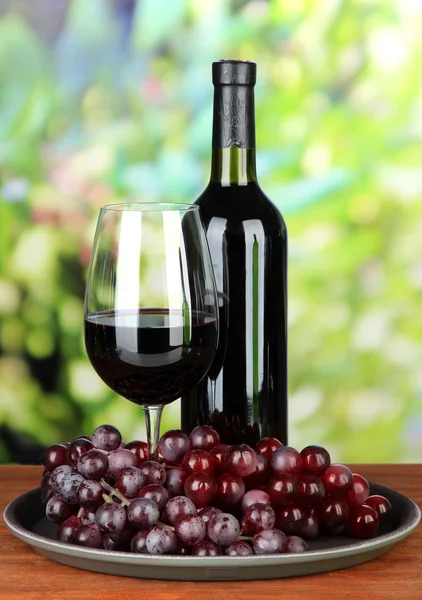 Uve mature, bottiglia e bicchiere di vino su vassoio, su sfondo brillante — Foto Stock