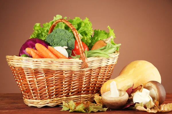Different vegetables in basket with yellow leaves on table on brown background — Stock Photo, Image