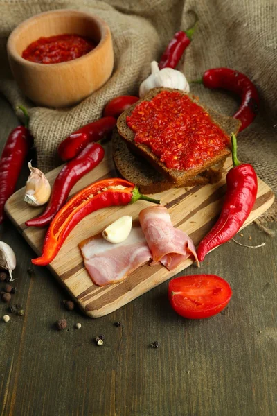 Composition with salsa sauce on bread,, red hot chili peppers and garlic, on sackcloth, on wooden background — Stock Photo, Image
