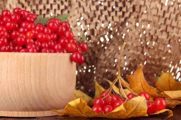 Red berries of viburnum in wooden bowl with yellow leaves on table on brown background — Stock Photo, Image