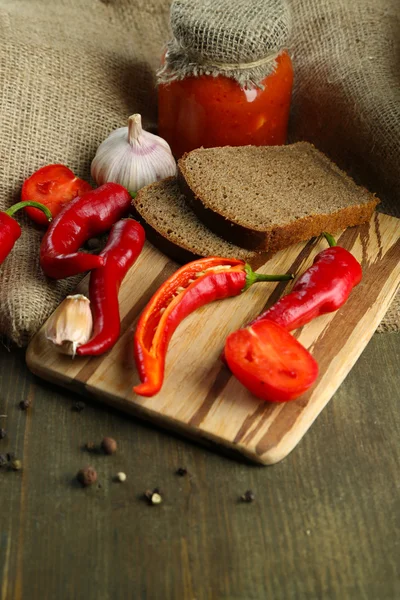 Composición con salsa en frasco de vidrio, pimientos rojos picantes y ajo, sobre tela de saco, sobre fondo de madera —  Fotos de Stock