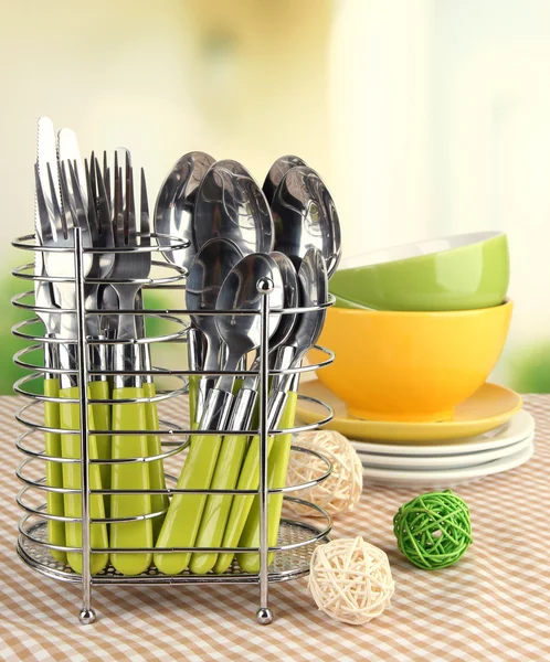 Kitchen cutlery in metal stand with clean dishes on tablecloth on bright background — Stock Photo, Image