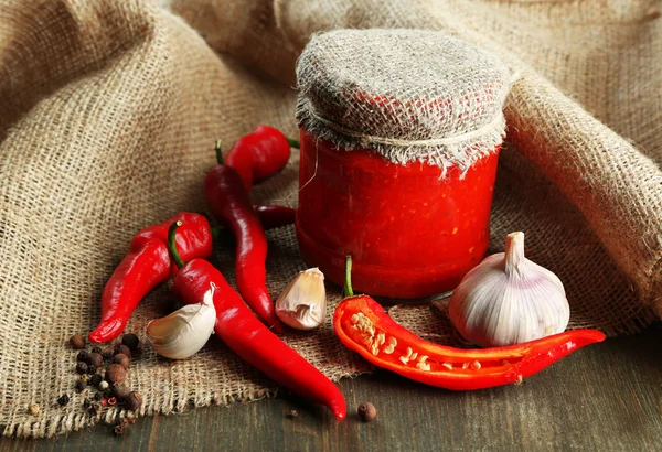 Composição com molho de salsa em jarra de vidro, pimenta vermelha e alho, sobre pano de saco, sobre fundo de madeira — Fotografia de Stock