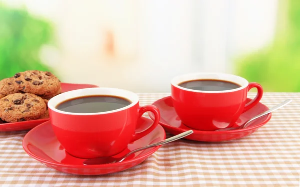 Copos vermelhos de café forte e biscoitos na toalha de mesa em fundo brilhante — Fotografia de Stock