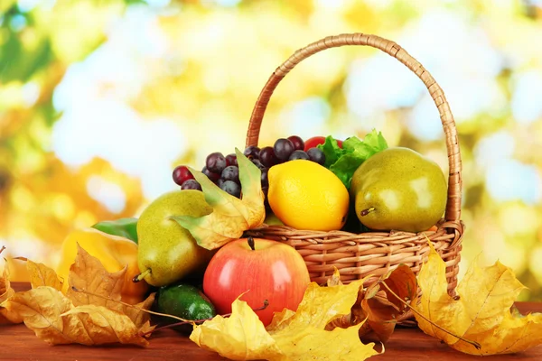 Diferentes frutas y verduras con hojas amarillas en canasta sobre mesa sobre fondo brillante — Foto de Stock