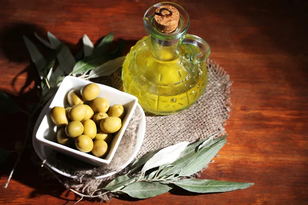 Aceite de oliva y aceitunas en tazón sobre tela de saco sobre mesa de madera — Foto de Stock