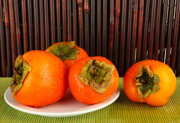Dojrzałe persimmons na talerz na stole na tle bambusa — Zdjęcie stockowe