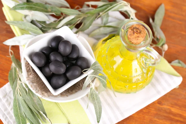 Olives in bowl and oil with branch on napkin on wooden board on table — Stock Photo, Image