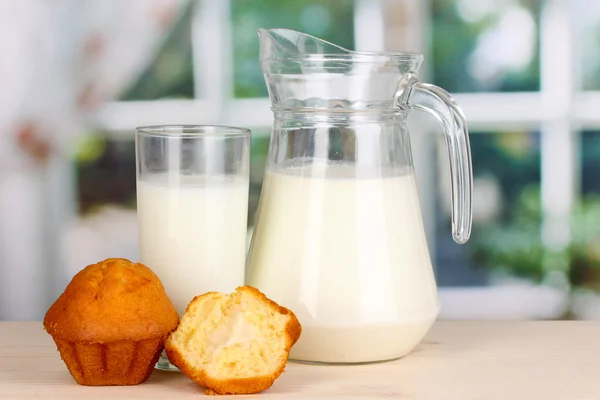 Jarra y vaso de leche con magdalenas sobre mesa de madera sobre fondo de ventana —  Fotos de Stock