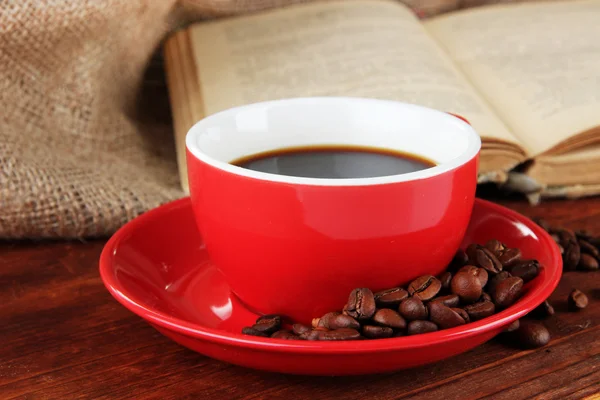 Чашка кофе с кофейными зёрнами и книга на деревянном столе на фоне мешковины — стоковое фото