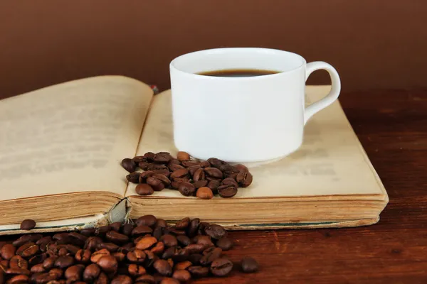 Cup of coffee with coffee beans and book on wooden table on brown background — Stock Photo, Image