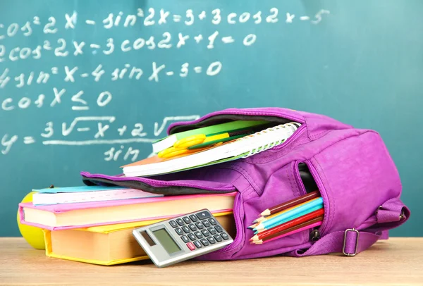 Purple backpack with school supplies on wooden table on green desk background — ストック写真