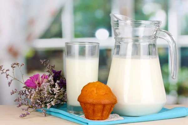 Jarro e copo de leite com muffins na mesa de madeira no fundo da janela — Fotografia de Stock