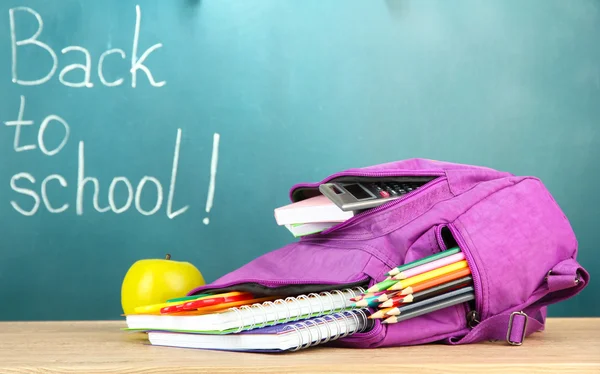 Purple backpack with school supplies on wooden table on green desk background — Stock Photo, Image