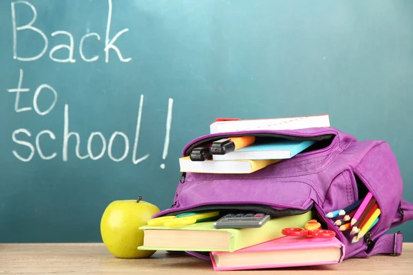 Purple backpack with school supplies on wooden table on green desk background — Stock Photo, Image