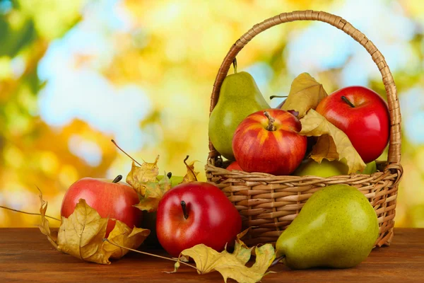 Hermosas manzanas maduras y peras con hojas amarillas en canasta sobre mesa sobre fondo brillante — Foto de Stock