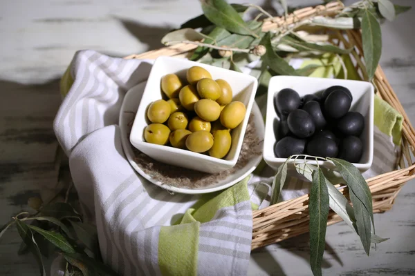 Olives in bowls with branch on napkin in basket on wooden table — Stock Photo, Image