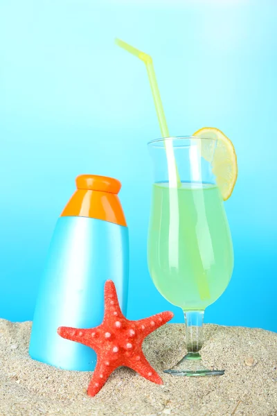 Strand cocktail en zonnebrandcrème in zand op blauwe achtergrond — Stockfoto