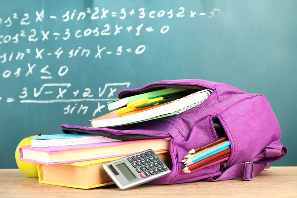 Purple backpack with school supplies on wooden table on green desk background — Stock Photo, Image