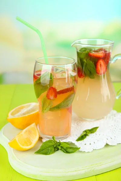 Basil lemonade with strawberry in jug and glass, on wooden table, on bright background — Stock Photo, Image