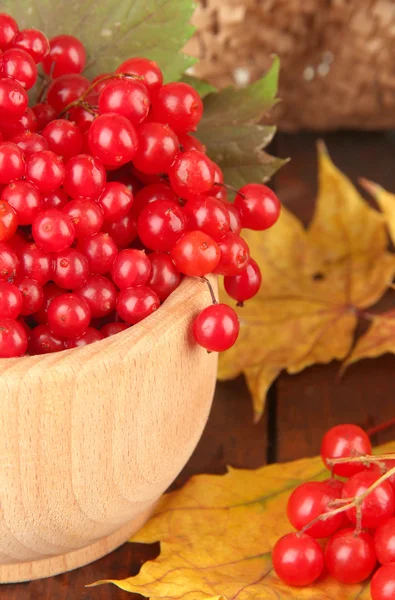 Czerwone jagody Kalina w drewniane miski z żółtymi liśćmi na stole na brązowym tle — Zdjęcie stockowe