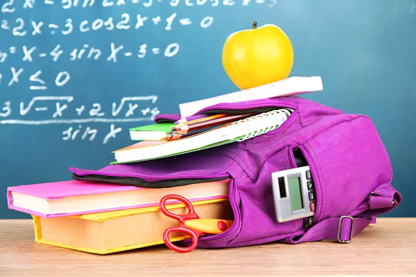 Фіолетовий рюкзак з шкільними приладдям на дерев'яному столі на фоні зеленого столу — стокове фото