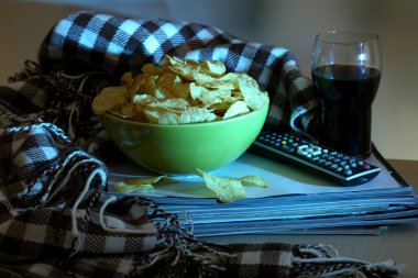 Chips in bowl, cola and TV remote on wooden table on room background clipart