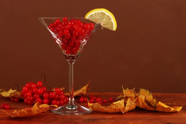 Red berries of viburnum in glass and yellow leaves on table on brown background — Stock Photo, Image