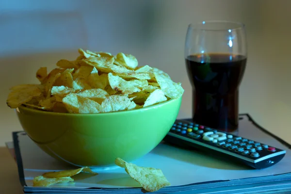 Chips in bowl, cola and TV remote on wooden table on room background — Stock Photo, Image